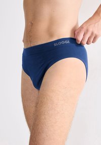 Mann, der blaue Sloggi-Unterhosen anpasst, von der Taille bis zur Mitte des Oberschenkels sichtbar, vor einem schlichten hellen Hintergrund.