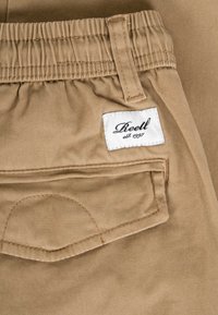 Nahaufnahme einer beigefarbenen Hosentasche mit elastischem Bund und einem weißen Stoffetikett mit der Aufschrift "Reell est. 1997."