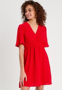 Robe rouge avec un décolleté en V, des manches courtes évasées et une taille élastique. Fabriquée en tissu léger, présentant une forme A-line fluide.