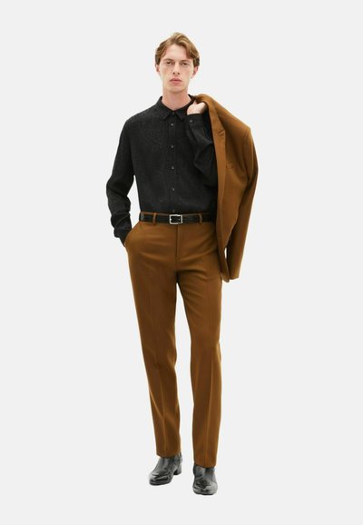 Zwarte geblokte overhemdd, bruine op maat gemaakte broek, zwarte riem en schoenen; persoon houdt een bruine blazer over één schouder.