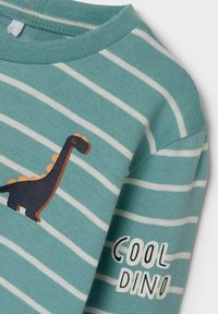 Turquoise gestreepte T-shirt van zachte stof met een zwarte dinosaurustekening en de tekst "COOL DINO" in het wit op de mouw.