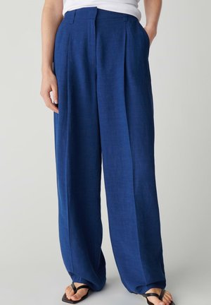 Persona che indossa pantaloni di lino blu larghi a vita alta abbinati a sandali neri aperti e una maglietta bianca.