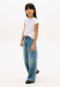 Weißes T-Shirt mit kurzen Ärmeln, kombiniert mit hellblauen, übergroßen Denim-Jeans und schwarzen klobigen Stiefeln, stehend vor einem weißen Hintergrund.