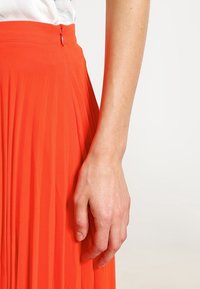 Plisovaná oranžová suknička s hladkou texturou, která má zipový uzávěr v pase. Materiál se zdá být lehký a splývavý.