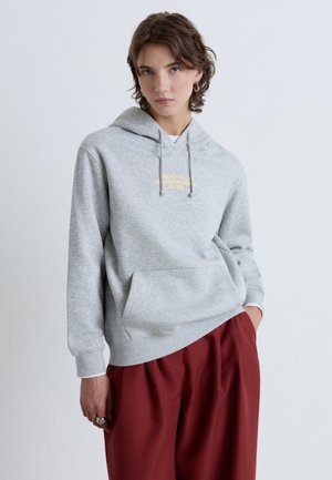 Personne aux cheveux courts et ondulés portant un sweat à capuche gris clair avec un texte jaune et un pantalon rouge ample, debout avec une main dans la poche.