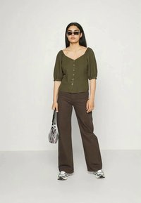 Blusa olive verde com mangas bufantes e botões, combinada com calças largas castanhas. Os acessórios incluem uma mala listrada em preto e branco e óculos de sol.