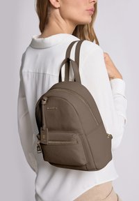 Mochila de cuero marrón con forma redondeada, dos correas para los hombros, cremalleras doradas, un bolsillo frontal y detalles texturizados.