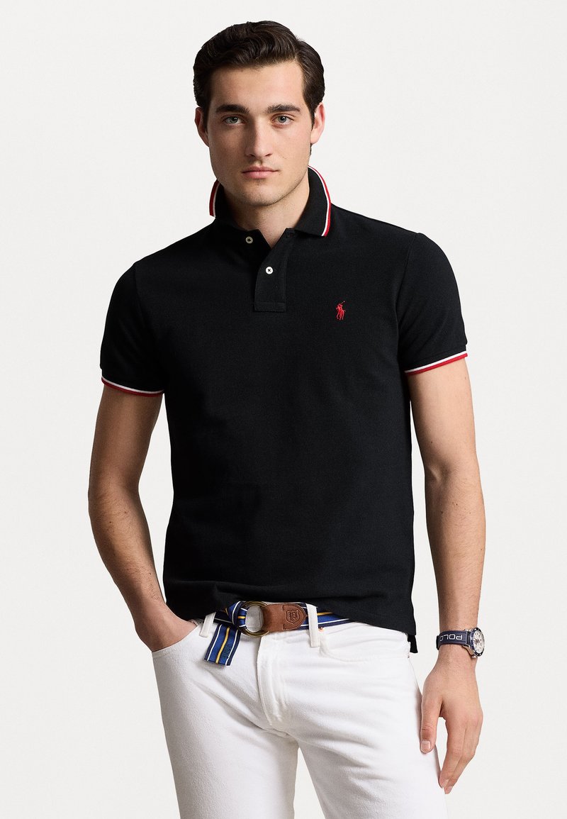 Black polo shirt with red and white striped collar, featuring a small red logo. Worn with white pants and a brown belt with blue accents.