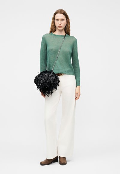Femme portant un pull vert et un pantalon blanc, tenant un sac à main noir texturé avec une bandoulière en chaîne, debout devant un fond uni.
