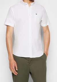 Biała koszula z krótkim rękawem, zapinana na guziki, z kieszonką, z małym haftowanym logo korony, wykonana z gładkiego materiału, w połączeniu z oliwkowymi spodniami.