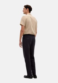 Beige Kurzarmhemd mit lockerer Passform, kombiniert mit dunkelblauen Hosen, die einen strukturierten Stoff und eine Gesäßtasche aufweisen.