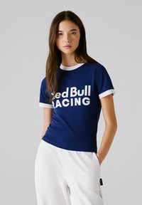 Giovane donna che indossa una maglietta blu scuro con il logo Red Bull Racing e bordi bianchi, pantaloni bianchi, in piedi davanti a uno sfondo grigio uniforme.