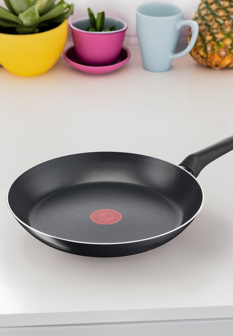 Sartén antiadherente negra con un punto indicador de calor rojo sobre una encimera blanca, rodeada de macetas de colores, una taza azul y una piña.