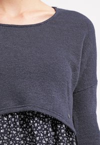 Mörkgrå, texturerad tröja med rund halsringning lager-på-lager över en svart klänning med blommönster. Har en kortad design och subtila hårdvaruaccent.