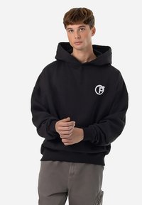 Schwarzer Kapuzenpullover mit lockerem Schnitt, versehen mit einem kleinen weißen Logo auf der Brust und gerippten Bündchen. Aus weichem Stoff gefertigt.
