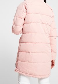 Žena v lehkém růžovém zimním kabátu, stojící zády k fotoaparátu, v bílých kalhotách.