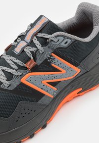 Černá sportovní bota s šedými mesh panely a oranžovými akcenty. Obsahuje vzorovanou vrstvu a strukturovanou podrážku pro lepší trakci. Šňůrky jsou šedé.