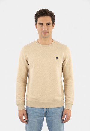 Uomo che indossa un maglione beige a maniche lunghe con un piccolo logo scuro a forma di albero sul petto e jeans azzurri, in piedi davanti a uno sfondo semplice.