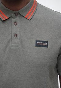 Κοντινό πλάνο ενός ατόμου που φοράει γκρι πόλο μπλούζα με κόκκινη ρίγα στο γιακά και ετικέτα της μάρκας Tom Tailor στο αριστερό στήθος.