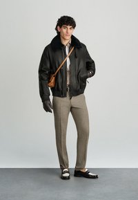 Černá kožená bomber bunda s kožichem, šedá košile na knoflíky, béžové kostkované kalhoty, černé rukavice a černobílé boty. Hnědá kabelka přes rameno.