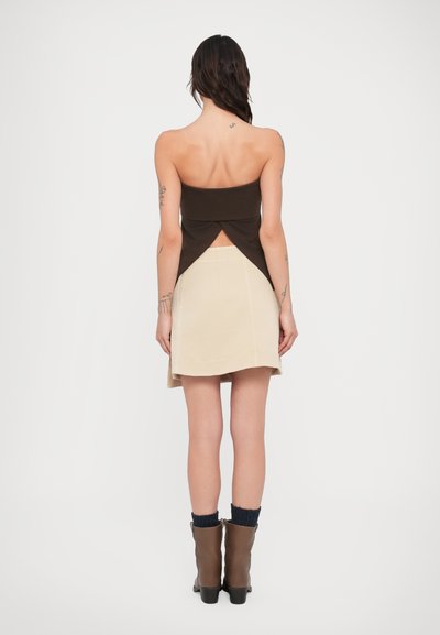 Καφέ τοπ χωρίς τιράντες με σχεδιασμό στριφογυρισμένης πλάτης, συνδυασμένο με φούστα κρεμ σε στυλ skater. Καφέ μποτάκια μέχρι τον αστράγαλο με πλεκτές λεπτομέρειες καλτσών.