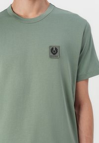 Tricou din bumbac verde, cu mâneci scurte, guler rotund și un mic patch cu logo negru pe piept. Textură netedă, croială casual.