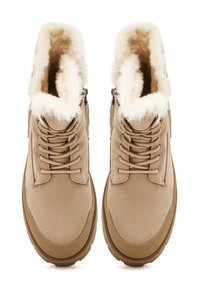 Paire de bottes d'hiver en daim beige avec semelles épaisses marron, lacets beige et doublure en polaire blanche et duveteuse visible à l'intérieur.