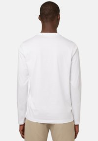 Valkoinen pitkähihaisella t-paidalla, jossa on klassinen pyöreä kaula-aukko. Pehmeä puuvillamateriaali, rento istuvuus ja sivuhelmoissa halkiot lisämukavuutta varten.