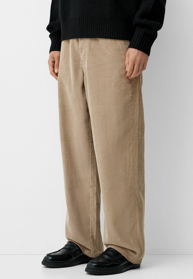 Personne portant un pantalon large beige en velours côtelé, un pull noir et des mocassins en cuir noir, debout devant un fond blanc.