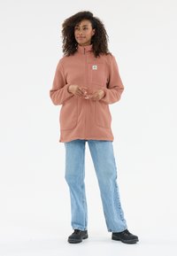 Fleecejacke in einem hellrosa Farbton, mit Reißverschluss vorne, hohem Kragen, einer Vordertasche und zwei Seitentaschen. Kombiniert mit hellblauen Jeans.