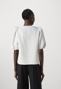 Donna con capelli trecciati che indossa una blusa bianca a maniche a sbuffo e pantaloni neri a gamba larga, in piedi contro uno sfondo chiaro e uniforme.