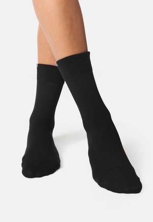 Chaussettes noires en coton à bord-côte, d'épaisseur moyenne, au design uni, portées sur un pied partiellement visible sur fond blanc.