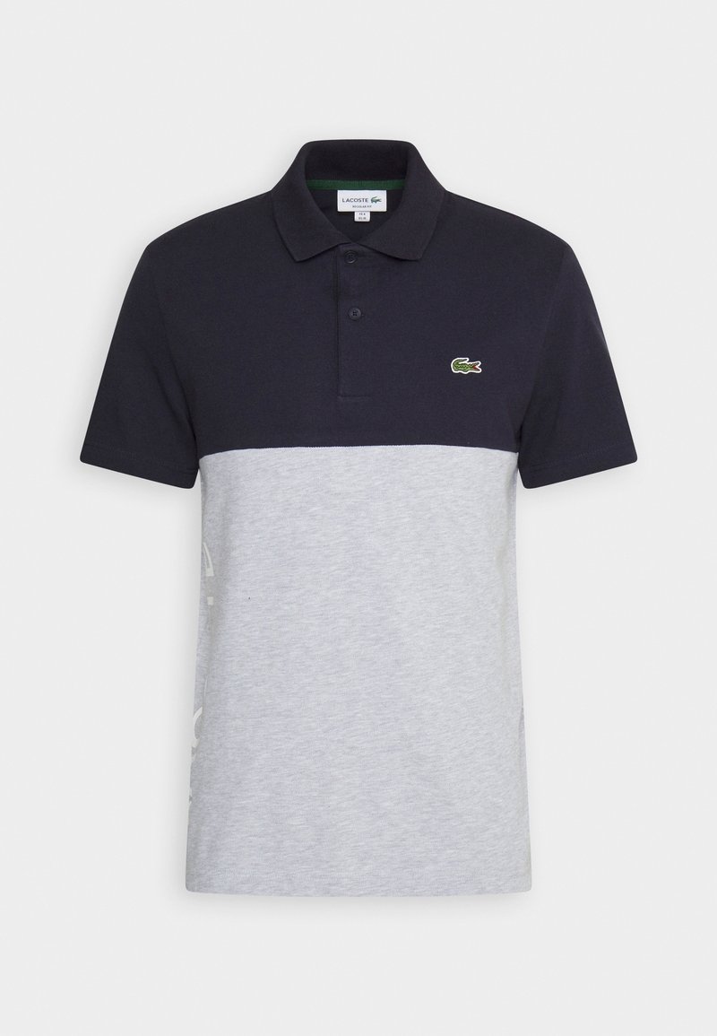 Polo à manches courtes avec moitié supérieure noire, moitié inférieure gris clair, deux boutons, col, et petit logo crocodile vert sur la poitrine.