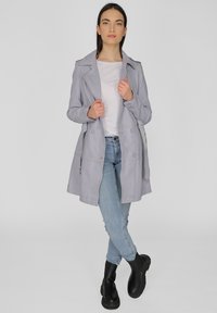 Lichtgrijze trenchcoat met revers, dubbele knopen sluiting en zijzakken, gecombineerd met lichtblauwe jeans en zwarte laarzen.