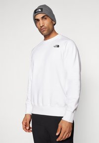 Witte sweatshirt met ronde hals, geribbelde manchetten en tailleband. Bevat een klein zwart logo op de linkerborst. Gecombineerd met een grijze beanie.