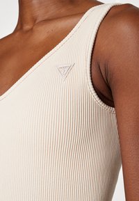 Beige ribbestrikket singlet med V-halsdesign, med et lite trekantet logoemblem på venstre side nær skulderen.