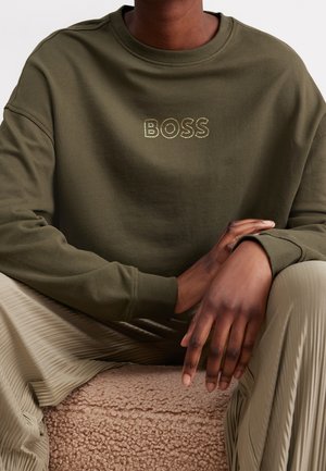 Person iført olivengrøn sweatshirt med "BOSS" broderet på brystet, siddende med hænderne hvilende på plisserede beige bukser.