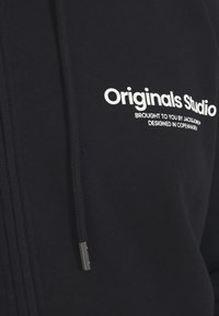 Marineblå hættetrøje med lynlås, der har et hvidt trykt logo "Originals Studio" og en snor med sølvspids. Blødt, struktureret materiale.