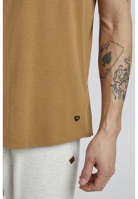 Brown cotton t-shirt with a rounded neckline and short sleeves, featuring a small black logo tag at the hem. Light gray sweatpants visible below.
