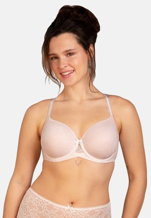 Smiling woman with dark hair in a light pink lace bra and matching underwear against a plain background.