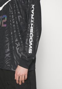 Chemise noire à manches longues avec un motif animal subtil, texte blanc "SWOOSH TRAX" sur la manche, et une partie du numéro irisé "72" sur la poitrine.
