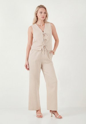 Frau trägt ein beigefarbenes, ärmelloses Oberteil mit drei vorderen Bändern, passende weite Hosen, Perlenkette und beige Sandaletten mit Absatz, steht vor weißem Hintergrund.