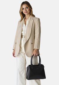 Borsa a tracolla nera in pelle con due manici superiori, forma rettangolare, superficie liscia e logo sottile sul davanti. Abbinata a un blazer beige e pantaloni chiari.