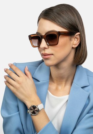 Brown square sunglasses with gradient lenses, paired with a black and silver watch on a white strap, worn by a person in a blue blazer.