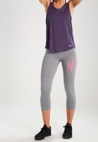 Lila ärmelloses Sport-Top mit Rundhalsausschnitt, kombiniert mit grauen Leggings bis zur Mitte der Waden, auf denen links "NIKE" in Pink steht. Schwarze Turnschuhe.