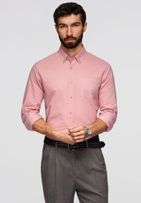 Homme aux cheveux foncés et à la barbe portant une chemise rose à boutons avec les manches retroussées, un pantalon gris, une ceinture noire et une montre-bracelet argentée, se tenant avec assurance.