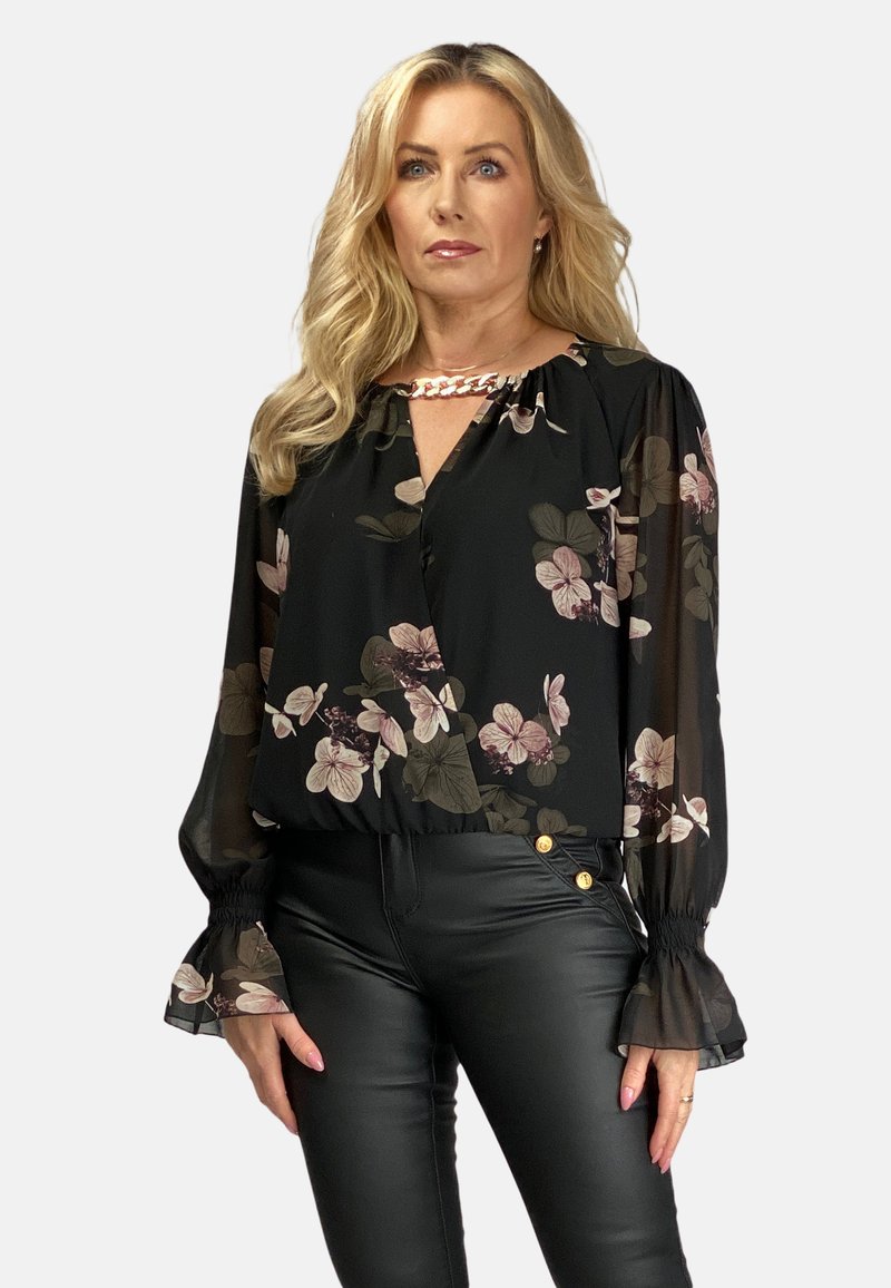 Czarna bluzka w kwiaty z przezroczystymi rękawami, z jasnoróżowymi i zielonymi wzorami kwiatowymi, zapinana na V, z elastycznymi mankietami. Połączona z czarnymi skórzanymi spodniami.