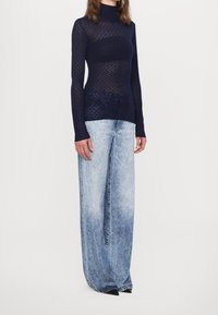 Maglia a maniche lunghe blu navy con collo a lupetto e motivo traforato geometrico, abbinata a jeans a gamba larga azzurri con finitura sbiadita.