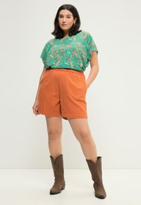 Zelený top s paisley vzorem a krátkými rukávy, doplněný o oranžové lněné šortky. Hnědé kovbojské boty outfit dokončují.