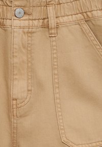 Beiger Denimstoff mit sichtbaren Nähten. Verfügt über einen elastischen Bund und zwei Vordertaschen, die durch einen markierten Metallknopf hervorgehoben werden.